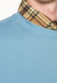Felpa azzurra con collo rotondo a coste, indossata sopra una camicia a quadri marrone e gialla con dettagli a bottoni.