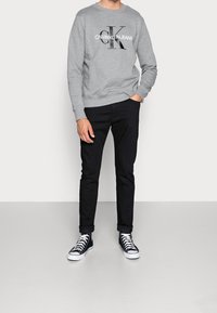 Šedá mikina s černým nápisem „CALVIN KLEIN JEANS“, doplněná černými úzkými džíny a černými kotníkovými teniskami s bílými podrážkami.