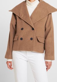 Brun dubbelknäppt ullkappa med stor krage, svarta knappar och sidofickor, bärs över en vit polo och byxor.