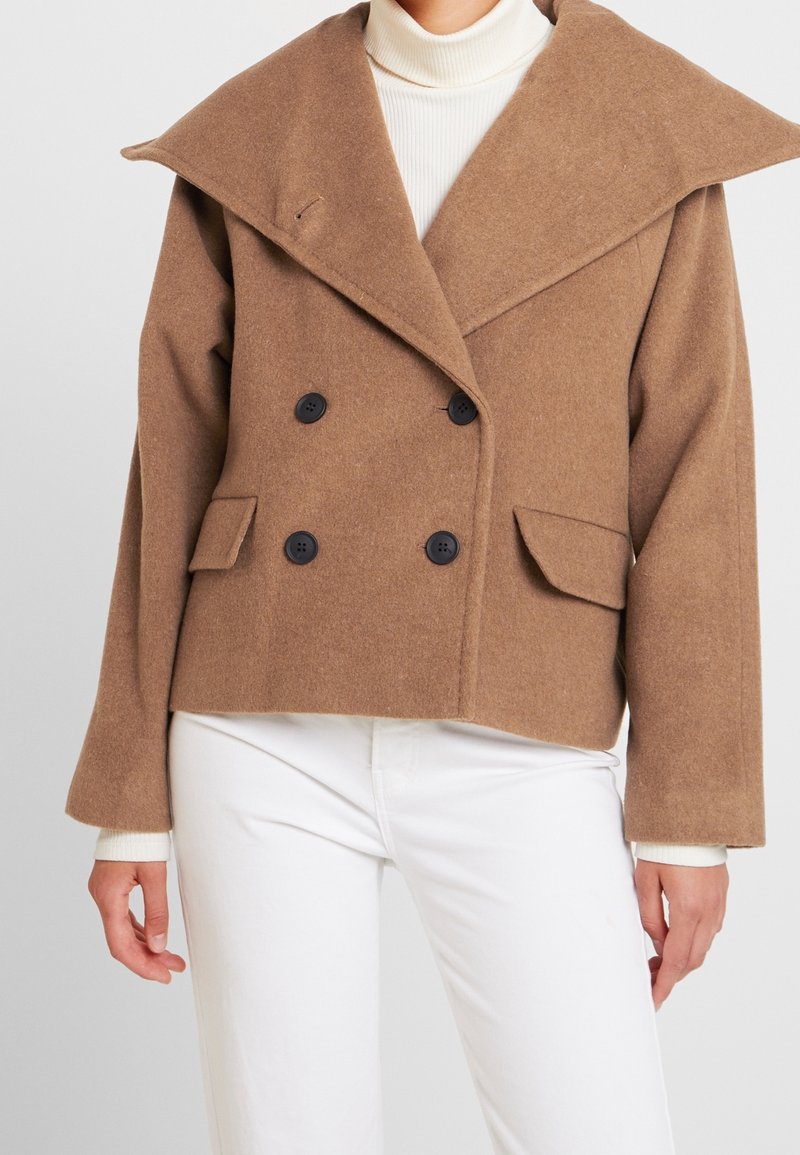 Brun dubbelknäppt ullkappa med stor krage, svarta knappar och sidofickor, bärs över en vit polo och byxor.