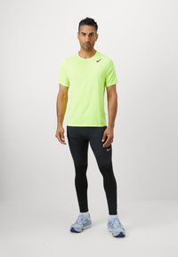Neon zielona koszulka sportowa z krótkim rękawem, w połączeniu z czarnymi legginsami i jasnoniebieskimi butami do biegania. Materiał wydaje się oddychający i teksturowany.