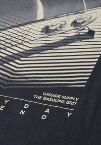 Gros plan sur le chevalet et les cordes d'une guitare, avec des lignes diagonales blanches et le texte "Garage Supply The Gasoline Grit" sur un fond en tissu sombre.