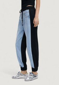 Pantalones deportivos de bloques de color con mezclilla azul claro y tejido negro. Cintura elástica con cordón. Tobillos ajustados. Zapatillas plateadas con superficie reflectante.