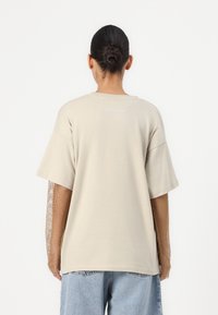Beige oversized t-shirt met een ronde hals en korte mouwen. Bevat mesh lange mouwen in een tan kleur. "H2O Fagerholt" logo op de achterkant.