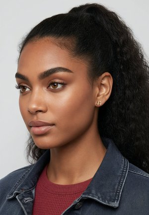 Boucles d'oreilles clous en ton or avec un design géométrique, portées par un mannequin aux cheveux frisés attachés en queue de cheval, sur un fond neutre.