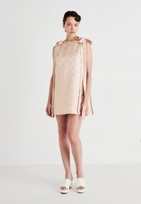 Frau, die in einem blassrosa ärmellosen Minikleid mit floralem Muster und langen Schleifen an den Schultern steht, trägt weiße offene Plateausandalen.