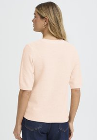 Gaiši rozā adīts džemperis ar īsām piedurknēm, apaļu izgriezumu un ribotām aprocēm, kopā valkāti ar tumši zilām džinsa biksēm.