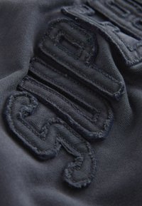 Tessuto scuro con lettering ricamato in rilievo. La texture appare morbida con una leggera lucentezza. I punti sono visibili lungo i bordi.