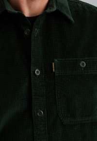 Donkergroen corduroy shirt met spitse kraag, knoopsluitingen en een borstzak. Beschikt over een ribbeltextuur en contrasterende knoopdetails.
