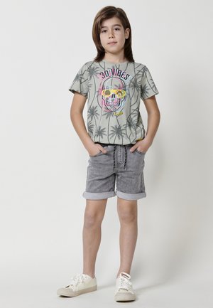 Maglietta grigia chiara con motivo di palme e grafica colorata di teschio, abbinata a pantaloni corti grigi con orlo arrotolato e sneakers bianche.