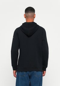 Uomo di spalle che indossa una felpa nera con cappuccio abbassato e jeans blu, su uno sfondo bianco uniforme.