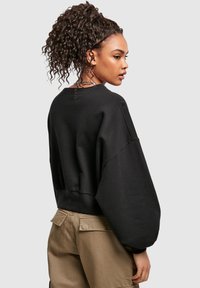 Kvinne med krøllete hestehale iført en svart oversized genser og beige bukser, som ser til siden mot en ensfarget grå bakgrunn.