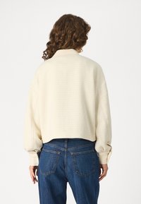Krämfärgad kort topp med strukturerad tyg, vida ärmar och hög halsringning, kombinerad med blå högmidjade jeans.