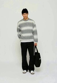 Jovem vestido com um suéter listrado em cinza e branco, gorro preto, calças pretas, tênis brancos, segurando uma bolsa preta contra um fundo simples.