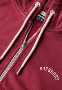 Sudadera con cremallera en burdeos, con cordones blancos, cremallera plateada y el logotipo "SUPERDRY" bordado en blanco. Textura suave con un diseño casual.