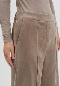 Gros plan sur une personne portant un pantalon en velours côtelé beige avec un haut à manches longues côtelé assorti, une main reposant sur leur côté.