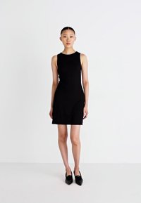 Czarna sukienka bez rękawów o dopasowanym kroju, długości do kolan i gładkiej teksturze. Połączona z czarnymi czółenkami na płaskim obcasie na jednolitym tle.