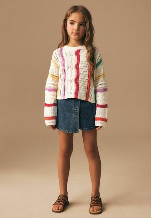 Young girl with long hair wearing a white knit sweater with colorful stripes, a denim skirt, and brown sandals standing against a beige background.
