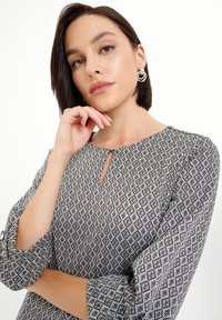Čiernobiele vzorované šaty s okrúhlym výstrihom, detailom v tvare kľúčovej dierky a trojštvrťovými rukávmi. Modelka má krátke tmavé vlasy a strieborné kruhové náušnice.