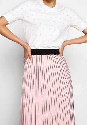 Femme portant une chemise blanche à pois manches courtes rentrée dans une jupe plissée rose avec une ceinture noire, les mains posées sur les hanches.