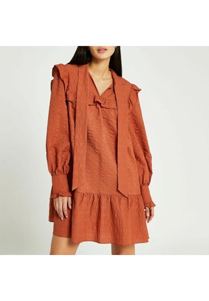 Femme portant une robe longue à manches longues texturée de couleur rouille-orange avec des détails à volants aux épaules et une jupe ample et évasée.