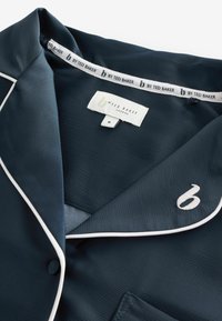 Camisa de pijama azul marino con ribete blanco, tela suave, cuello con muesca, detalle bordado con logo y un solo bolsillo. Etiqueta de talla visible.