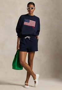 Ženska, oblečena v mornarsko modro majico s kapuco z ameriško zastavo, ujemajoče kratke hlače, bele čevlje in sončna očala, drži zeleno torbo za na ramo.