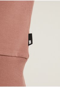 Gros plan sur l'ourlet d'un pull côtelé rose poussiéreux avec une petite étiquette en tissu noire et blanche sur le côté, sur un fond clair.