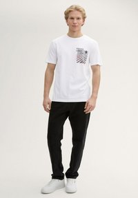 Weißes T-Shirt mit kurzen Ärmeln und grafischem Druck auf der linken Brust. Kombiniert mit schwarzen Jeans und weißen Sneakers. Glatte Textur, normale Passform.