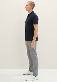 Navyblauwe polo met korte mouwen en een kraag, gecombineerd met lichtgrijze broek en witte sneakers, gepresenteerd vanuit een zij-aanzicht.