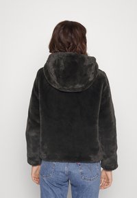 Chaqueta de pelaje sintético negra con una capucha amplia; textura suave y corte relajado. Combinada con unos pantalones vaqueros azules visibles en la parte inferior.