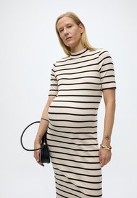 Έγκυος γυναίκα που φοράει μπεζ και μαύρο ριγέ φόρεμα, κρατώντας μια μαύρη τσάντα, με χρυσό βραχιόλι και μικρά τατουάζ στα πήχεις.