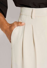 Roomkleurige geplooide broek met een aangesloten taille, voorzien van zijzakken en een gladde, zachte stoftextuur.