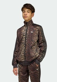 Hnedo-čierna pruhovaná tepláková súprava so zipsovou bundou a nohavicami v rovnakej farbe. Na rukávoch sú tri čierne pruhy, na hrudi logo.