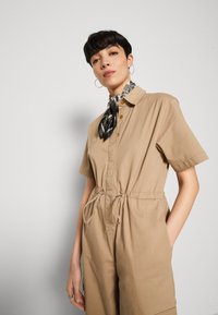 Khaki short-sleeve jumpsuit with a buttoned collar, drawstring waist, and side pockets; features a black and white patterned scarf tied at neck.
