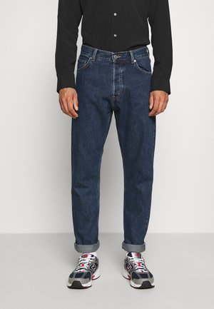 Jeans de mezclilla azul oscuro con un corte recto, que cuentan con un cierre de botón y cremallera, cinco bolsillos y un ligero deslavado. Combinados con zapatillas blancas y grises.