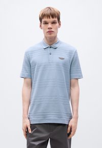 Ανοιχτό μπλε  πόλο με οριζόντιες ρίγες, κοντά μανίκια και γιακά με κουμπιά. Διαθέτει μικρό καφέ λογότυπο στο στήθος.