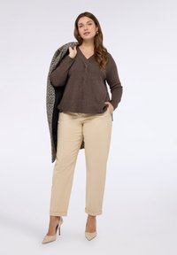 Kobieta w brązowym swetrze w serek i beżowych spodniach, trzymająca teksturowany płaszcz na ramieniu, stojąca na jednolitym tle.