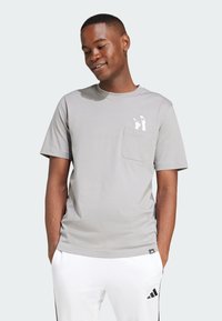 Šedé tričko s krátkým rukávem a kulatým výstřihem, které má kapsu na levé straně hrudi a bílý potisk. Hladká textura, volný střih.