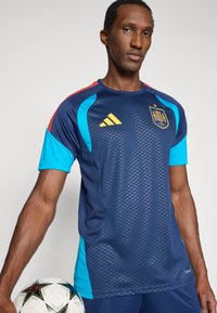 Vīrietis, kurš valkā zilu Spānijas futbola kreklu, tur baltu futbola bumbu ar melniem zvaigžņu rakstiem uz vienkārša fona.