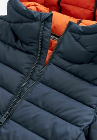Veste en duvet navy avec un intérieur orange, dotée d'une fermeture éclair sur toute la longueur et d'un design matelassé pour l'isolation et la chaleur.