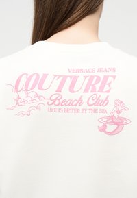 Λευκό πίσω μέρος πουκαμίσου με ροζ κείμενο "Versace Jeans Couture Beach Club" και εικόνα γοργόνας, με στυλιζαρισμένα σύννεφα και τη φράση "Η ζωή είναι καλύτερη δίπλα στη θάλασσα."