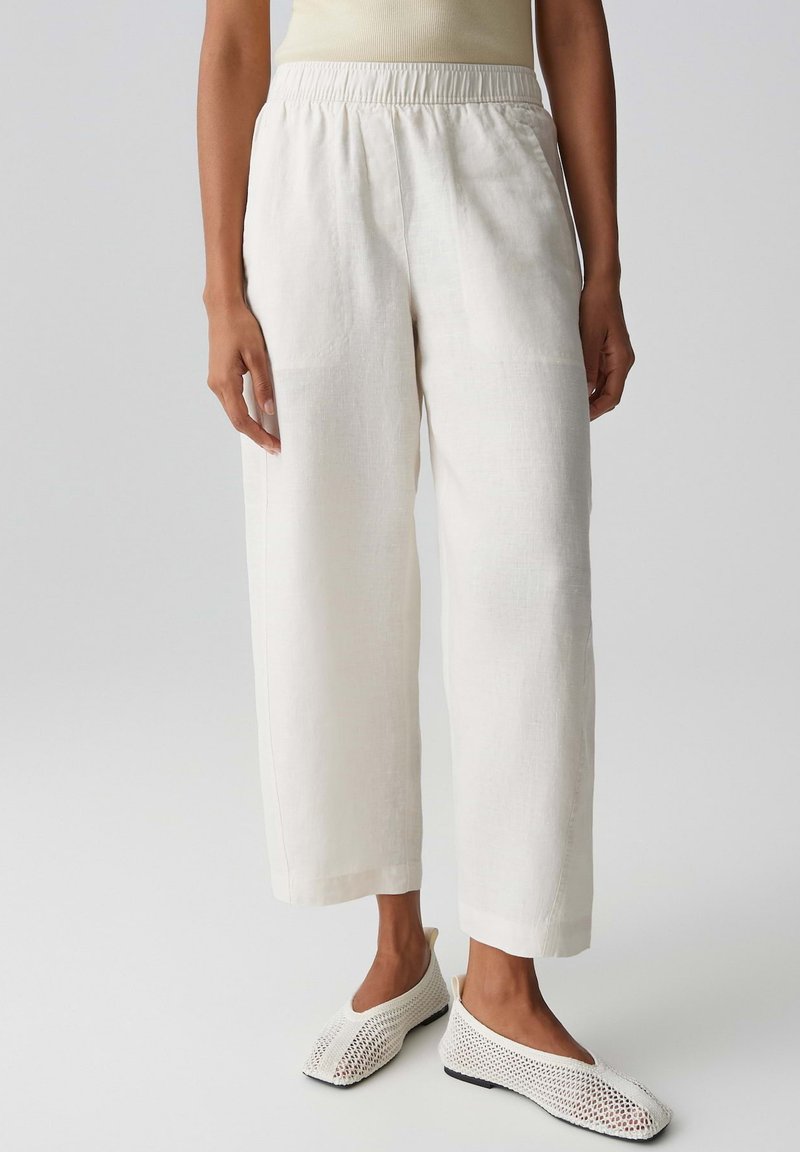 Person wearing loose white ankle-length pants and white mesh slip-on shoes, standing on a plain light gray background.