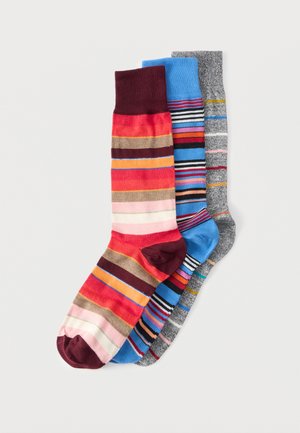 Trois paires de chaussettes rayées colorées, une ton rouge, une ton bleu et une ton gris, disposées en chevauchement sur un fond blanc.