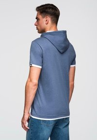 Krátký rukáv modré mikiny s bílým lemem na rukávech, se stahovací kapucí a rovný střih, vyrobené z měkkého materiálu.