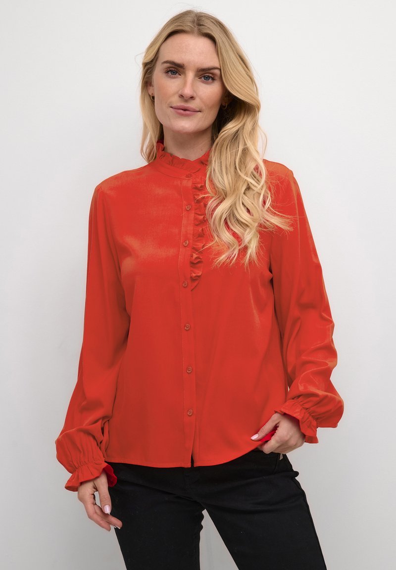 Red satin blouse with ruffled collar and front, long sleeves with gathered cuffs, and button-down front. Paired with black pants.