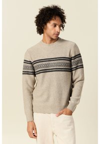 Suéter de punto beige con cuello redondo, mangas largas y una banda negra con patrón en el pecho. Textura suave, puños acanalados.