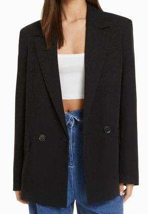 Femme portant un blazer noir oversize croisé sur un crop top blanc et un jean bleu taille haute, debout sur fond blanc.