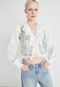 Gilet cropped in denim argentato metallizzato con colletto volant sopra una camicia bianca. Abbinato a jeans blu chiarissimo con vestibilità classica.