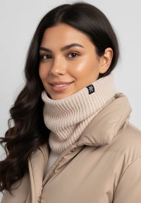 Beige knitted scarf with a ribbed texture and black tag, worn with a beige puffer jacket. Scarf is positioned around the neck.